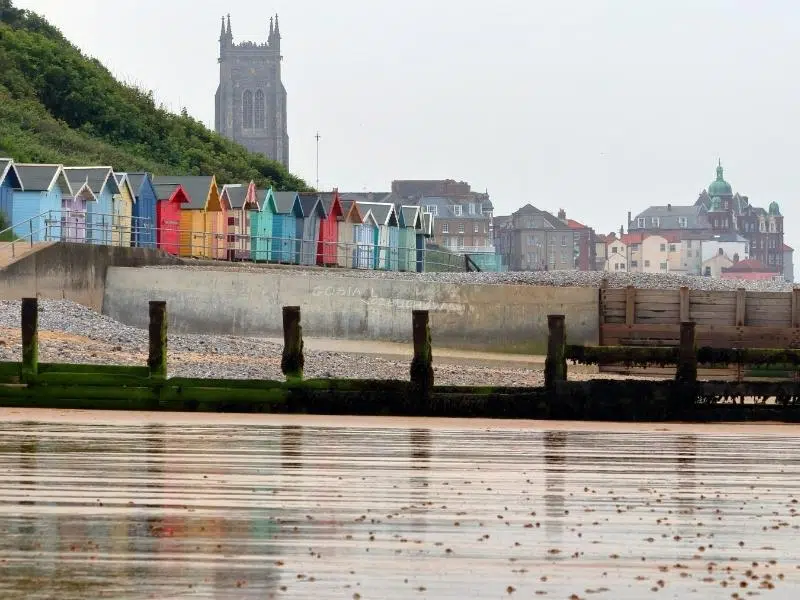 A row of colourful wooden beach huts facing a sandy beach with a town and church tower in the background