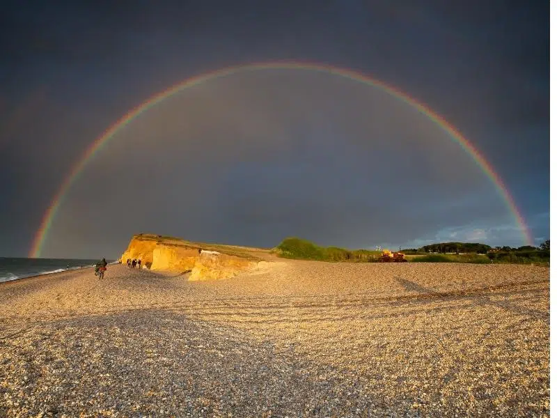 a beach with orange cliffs and a full rainbow above against a dark sky