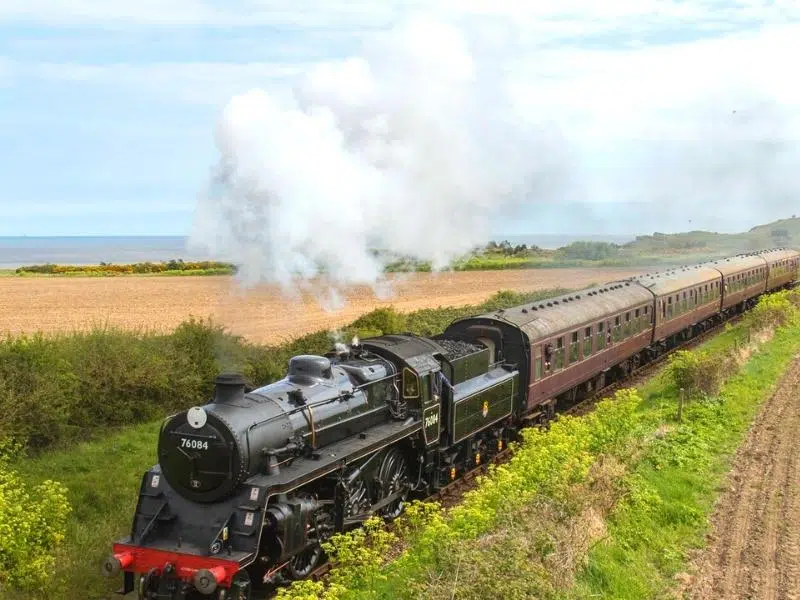 a steam train blowing steam between green vegetation, wheat fields and sea
