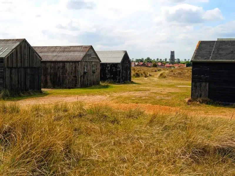 black fishermans huts amongst grasses and dunes with a church and village in the background