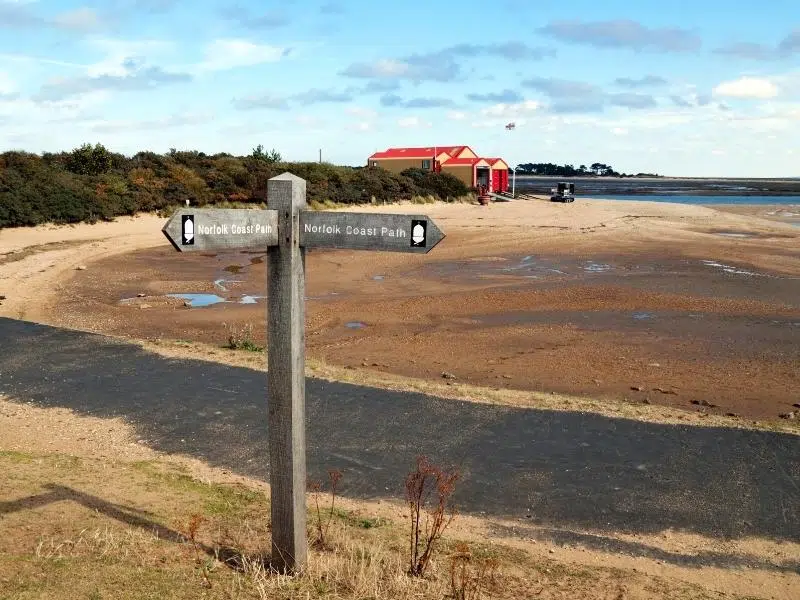 Norfolk coastal path sign post next to a beach with red and cream shed in the background