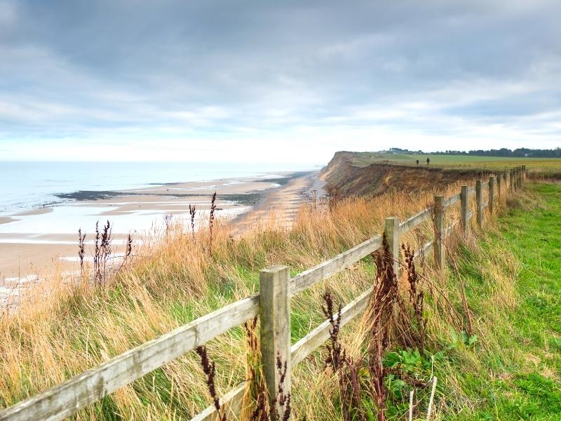 view across wooden fence and grasses to a sandy beach with tide partially out, backed by small cliffs