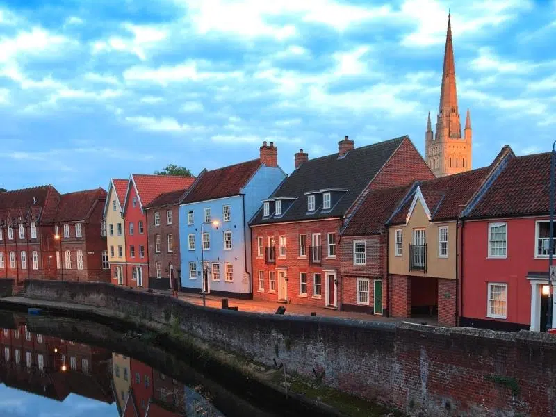 row of colourful historic houses along a river, with a catherdal spire in the distance