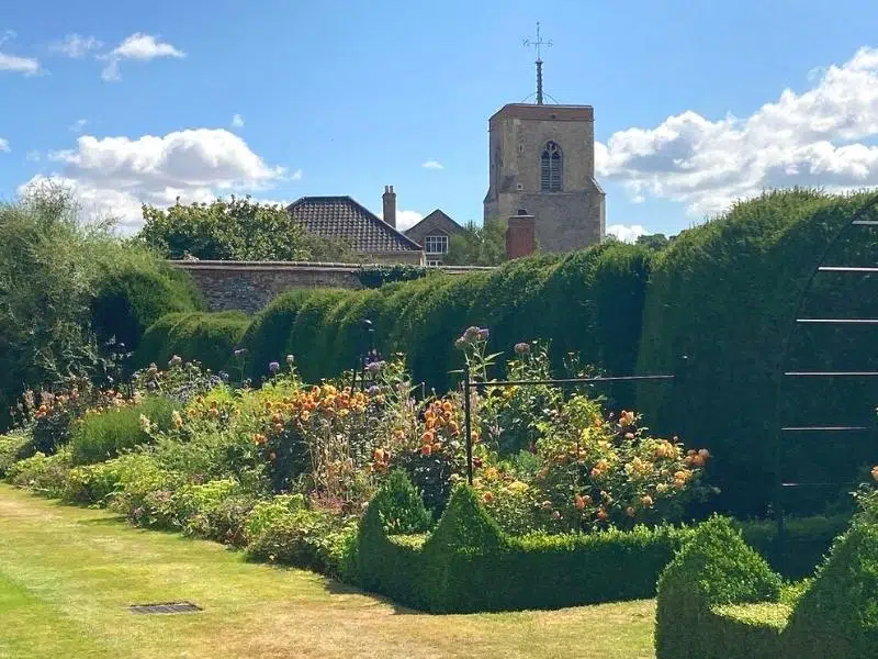tall green hedging with a church tower in the background and flower beds and a lawn in the foreground
