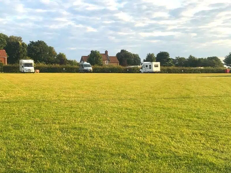 Motorhomes and a campervan parked in a large grass field by a hedge and house