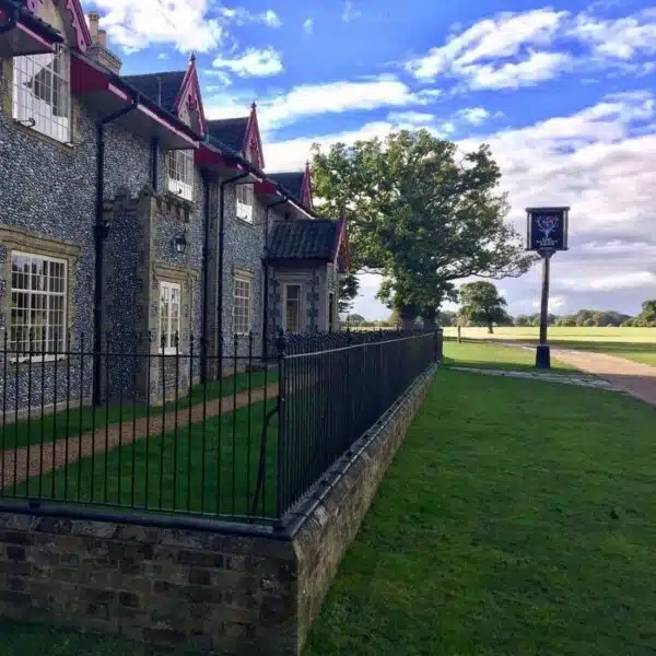 An image of a stone and flint building behind a wrought iron low fence, with a blue sky and tree in the background and a pub sign