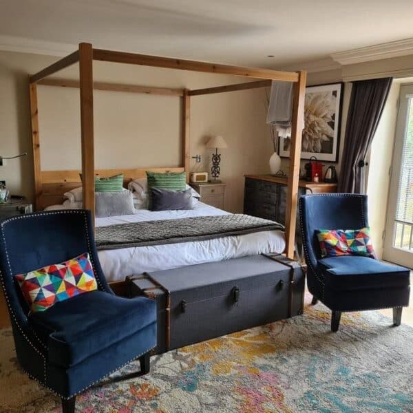 An image of a hotel room, with a modern 4 poster bed, with two blue velvet chairs with multicoloured cushions and a pastel coloured rug