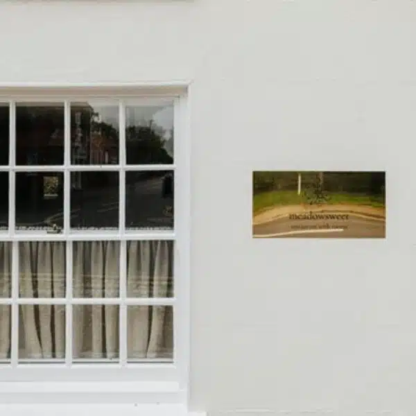 An white wall with a partial sash window and a gold plaque engraved in black saying Meadowsweet