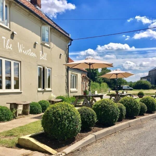 An image of the front of a building painted sage green, with the words The Wiveton Bell painted in white cursive. there are tables with Umbrellas and round bushes in the front.