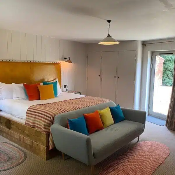 An image of a hotel room with a wooden bed, with a tan leather headboard and grey sofa and red, yellow and blue cushions