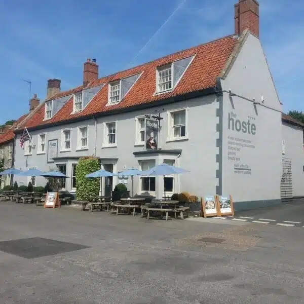 An image of a grey pub, with a red roof and tables and blue umbrellas outside, on the side of the building is written The Hoste