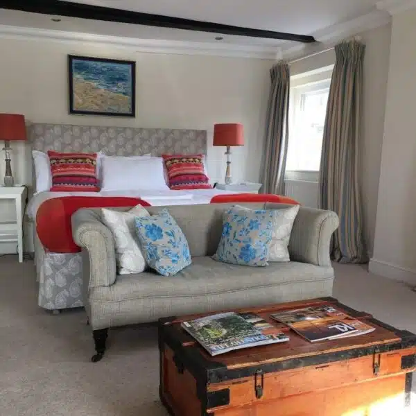 An image of a hotels room with grey bed and sofa with red and blue cushions and a wooden trunk in front of the bed