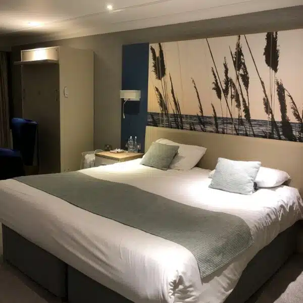 An image of a double bed with white sheets a grey runner at the bottom, there is a black and white painting of pampas grass behind the bed.