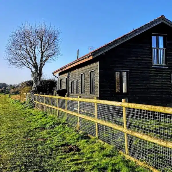 An image of the side of a large back barn, in a filed with metal and wood fence and a tree in the background.