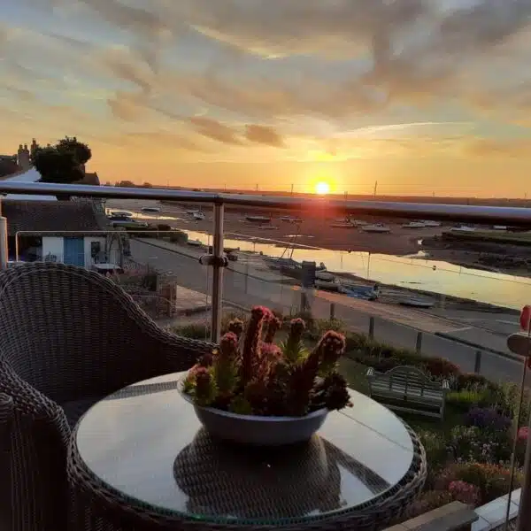 An image of a glass table with a wicker chair with a plant on top, looking over the harbour with some boats at low tide.