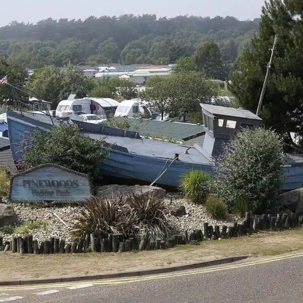 An image of the entrance to a campsite, with a sign saying Pinewoods Holiday Park and a an old blue wooden boat.