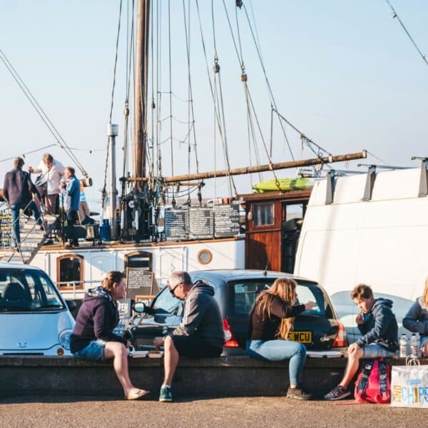 people eating fish and chips on a harbour wall with a sailing boat and cars beyond