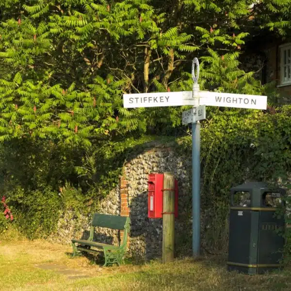 Traditional road sign post by a red postbox and a green bench, surrounded by green foliage and trees