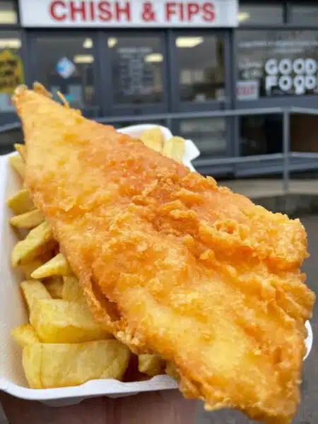 Crispy battered cod and chips in a white platic tray, in front of a chip shop