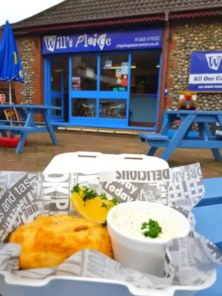 Fish and chips with a white pot of tartare sauce, served in newspaper and a white plastic tray on a blue wooden table outside a chippy