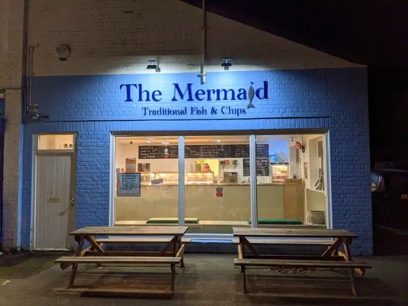 Blue painted fish bar in the evening, with wooden picnic tables out front
