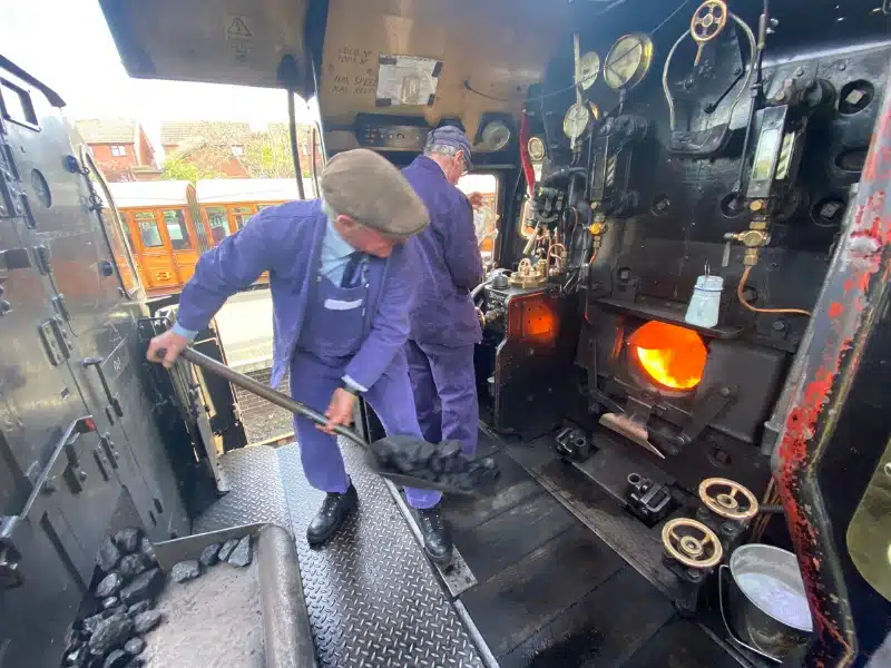 Stoker in a flat cap and blue uniform shovelling coal into a steam engine