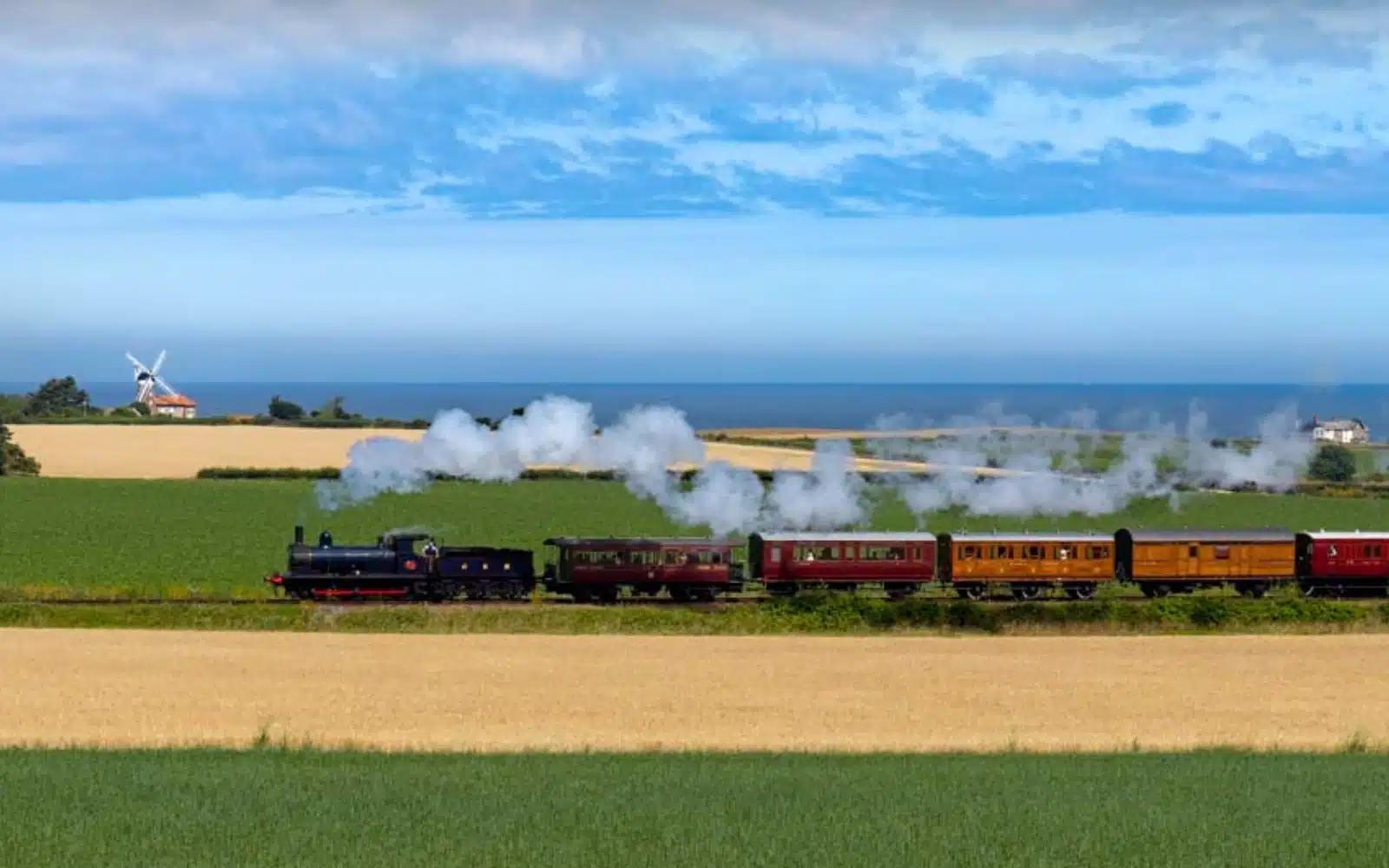 Steam train puffing across green and yellow fields with a windmill and the sea in the background 