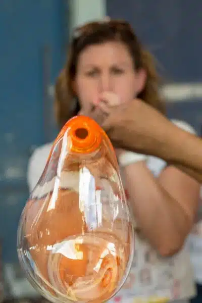 lady blowing an orange glass bubble through a long pipe