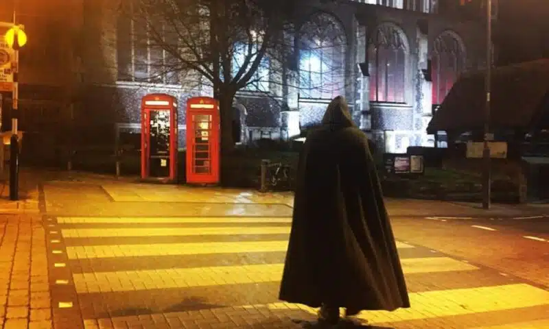 person in a hooded cape crossing a road at night with two red phone boxes in front of a church
