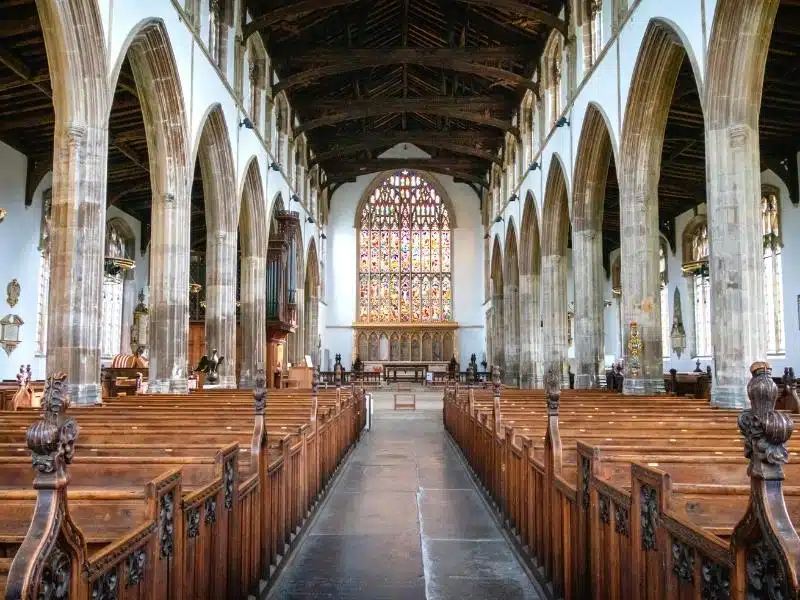 Large chapel with seating and pillars, and a stained glass window at the end of the aisle